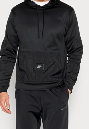 Muž nosící černou mikinu Nike s přední kapsou a odpovídající černé kalhoty Nike, ruce volně podél těla, proti jednoduchému pozadí.