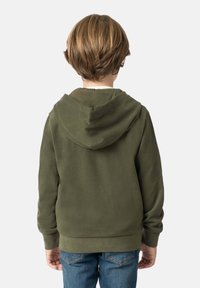 Felpa verde oliva con una texture morbida, caratterizzata da un grande cappuccio e polsini a coste. Indossata sopra una camicia chiara e abbinata a jeans blu.