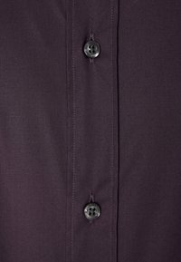 Gros plan sur un tissu violet foncé avec deux boutons noirs cousus verticalement le long d’une patte de boutonnage cousue.