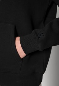 Un primer plano del brazo de una persona que lleva un sudadera negra con bolsillo, mostrando una mano descansando en el bolsillo y el puño con un logo.