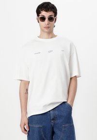 Übergroßes weißes Baumwoll-T-Shirt mit kurzen Ärmeln, mit lila Logodetails auf der Vorderseite und einem Rundhalsausschnitt.