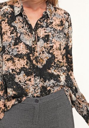 Femme portant une chemise à manches longues avec des boutons, ornée d'un motif abstrait noir, pêche et gris, associée à un pantalon gris taille haute.