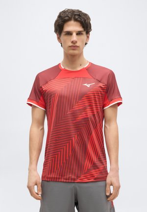 Νέος άνδρας που στέκεται φορώντας κόκκινο αθλητικό μπλουζάκι με γεωμετρικές ρίγες και λευκό λογότυπο, συνδυασμένο με γκρι σορτς, ουδέτερο φόντο.