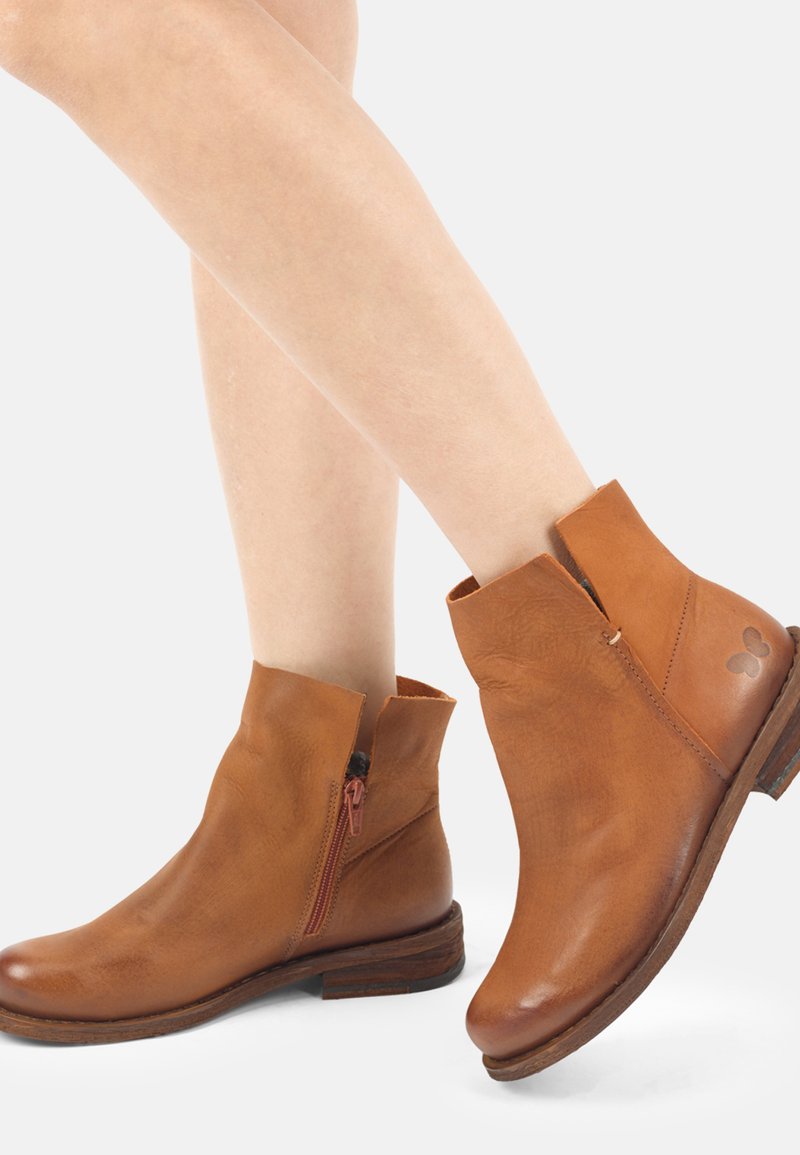 Gros plan sur des jambes portant des bottines en cuir beige avec des fermetures éclair latérales et des talons bas en bois sur un fond clair.