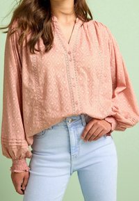 Blusa rosa chiaro, con un motivo a diamante testurizzato, chiusura con bottoni frontale e maniche a sbuffo con polsini arricciati. Abbinata a jeans azzurri.