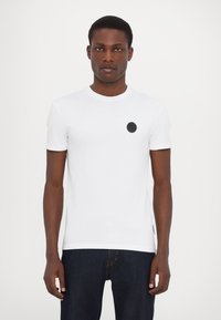Hvid kortærmet t-shirt i blød bomuld, med et sort cirkulært logo på venstre bryst og rund halsudskæring. Kombination med mørke jeans.