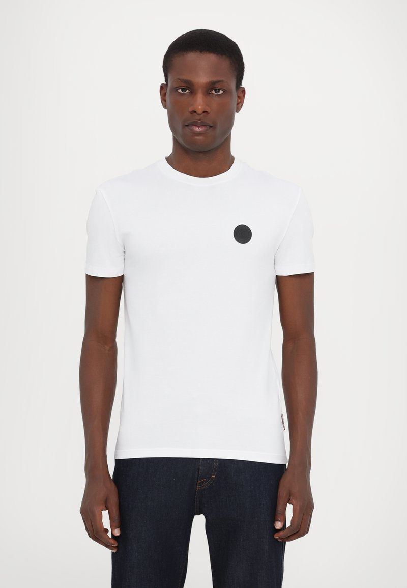 Hvid kortærmet t-shirt i blød bomuld, med et sort cirkulært logo på venstre bryst og rund halsudskæring. Kombination med mørke jeans.