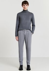 Grauer Rollkragenpullover kombiniert mit hellgrauen Hosen aus strukturiertem Stoff, mit Falten und einem Gürtel. Das Model trägt schwarze Stiefeletten.