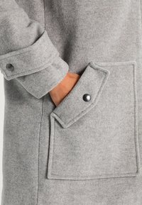 Grå uldfrakke med stor påsat lomme med klap, knaplukning på manchet og blød tekstur.