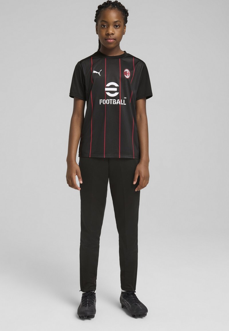 Zwart voetbalshirt met rode strepen, met Puma-logo en de tekst 'FOOTBALL'. Gecombineerd met zwarte broek en zwarte sportschoenen.