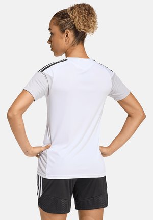 Kobieta o kręconych włosach w białej sportowej koszulce z krótkim rękawem i czarnych szortach, stojąca z rękami na biodrach, zwrócona tyłem.