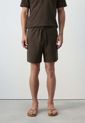 Hombre con pantalones cortos marrones, camiseta marrón a juego y sandalias color beige, de pie sobre un suelo gris frente a una pared blanca.