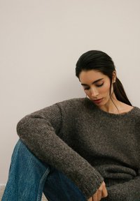 Przesadnie duży brązowy sweter dzianinowy o teksturowanym wykończeniu i szerokich rękawach, w zestawieniu z niebieskimi dżinsami. Prosty, luźny krój.