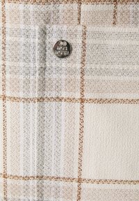 Tissu texturé présentant un motif à carreaux crème et gris clair avec des accents marron, complété par un bouton logo rond en métal.