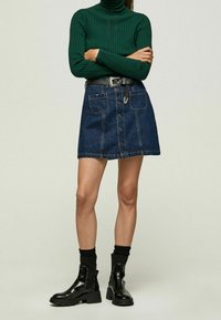 Personne portant un pull à col roulé côtelé vert foncé, une jupe en denim mini boutonnée avec des poches avant, une ceinture noire, des chaussettes noires et des bottines noires brillantes.