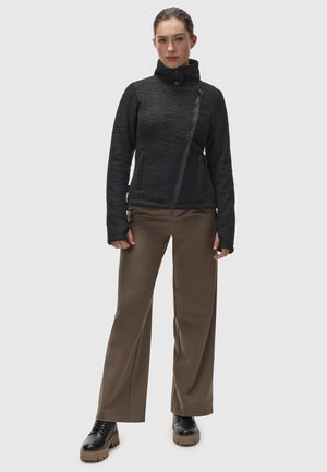 Schwarze Jacke mit Reißverschluss und hohem Kragen sowie strukturiertem Finish, kombiniert mit braunen Hosen im weiten Schnitt und schwarzen Schnürstiefeln auf hellem Hintergrund.
