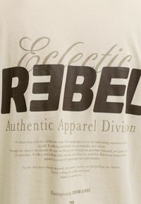 Tissu beige avec un grand texte en gras "REBEL", "Eclectic" en cursive, et un texte plus petit sur les vêtements exprimant l'individualité et la personnalité.