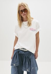 Lyshåret person med langt hår iført store brune solbriller, hvid T-shirt med op rullede ærmer og pink tekst, denimjakke bundet om livet og jeans.