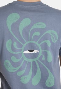 Camiseta de algodón azul con un patrón de espirales verdes y un diseño de ojo central en blanco y negro en la parte posterior.