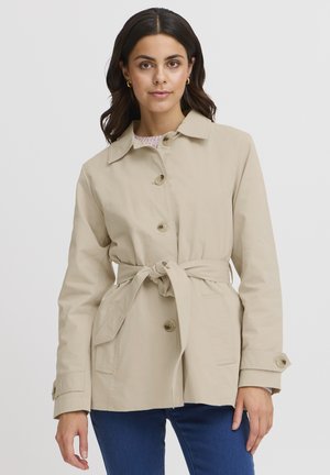 Jachetă bej cu curea, guler cu rever, închidere cu nasturi, două buzunare frontale și o textură netedă a țesăturii.