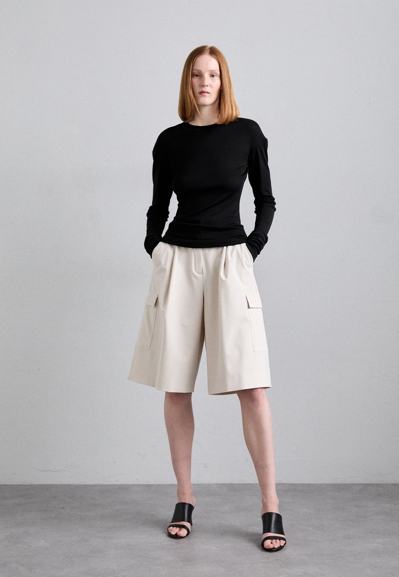 Schwarzes Langarmoberteil, beigefarbene weite Shorts mit zwei vorderen Taschen und schwarze Slipper-Sandalen. Glatte Texturen, einfaches Design, neutrale Farben.