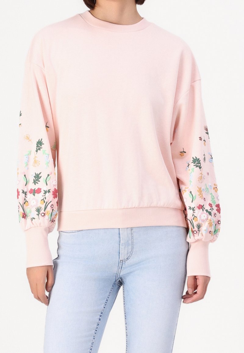 Femme portant un sweat-shirt rose clair avec une broderie florale colorée sur les manches bouffantes et un jean bleu clair, debout devant un fond blanc.