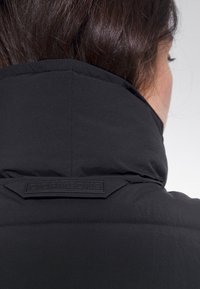 Svart vadderad jacka med hög krage och präglad logotags vid halsringningen, med en slät, matt yta.