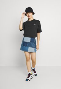 T-shirt nero oversize, minigonna di jeans con toppe blu miste e sneakers sportive colorate. Gli accessori includono un cappello a secchiello nero.