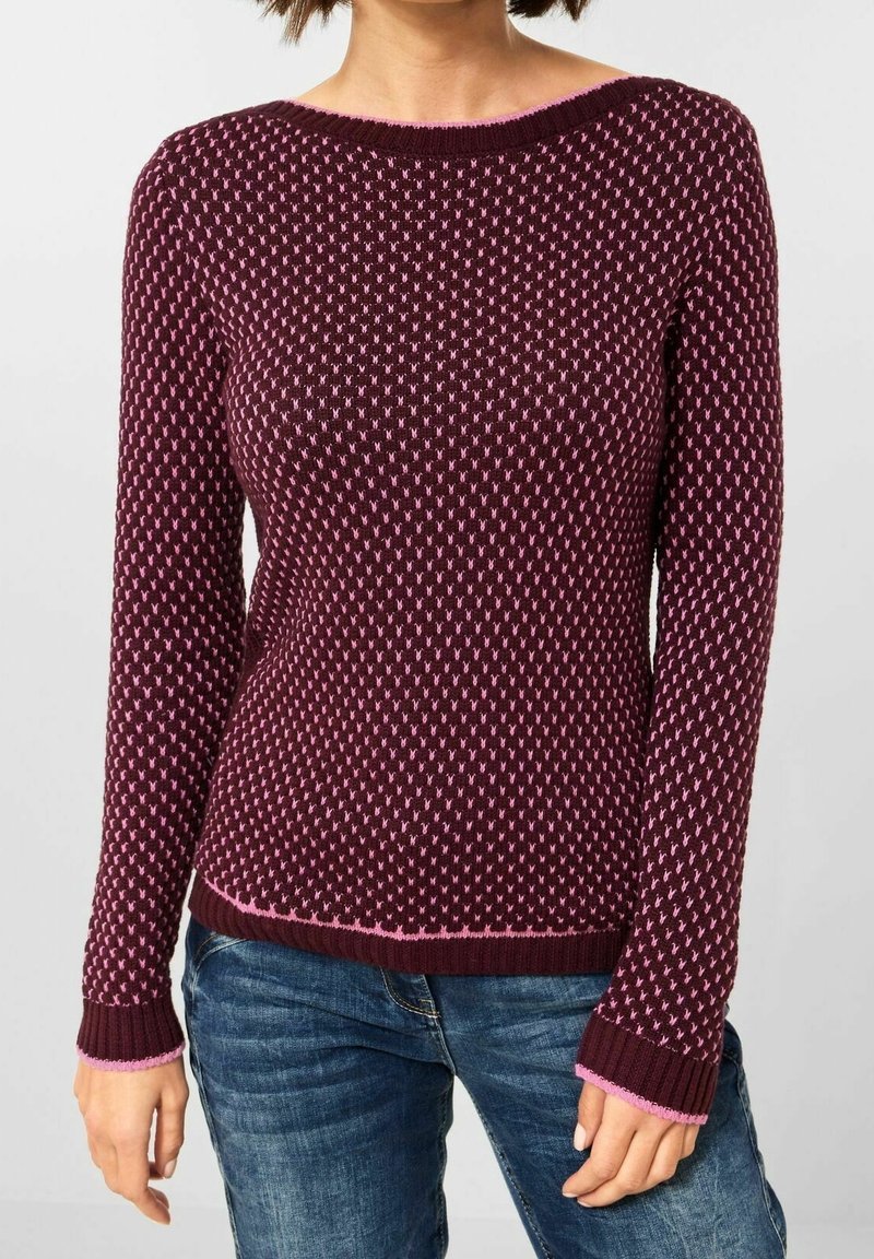 Femme portant un pull en maille ajusté bordeaux avec un motif à pois roses et un jean bleu, debout devant un fond clair uni.