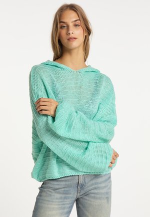 Ung kvinde iført en løs mintgrøn strikket trøje med hætte og lyseblå jeans, står med armene over kors.