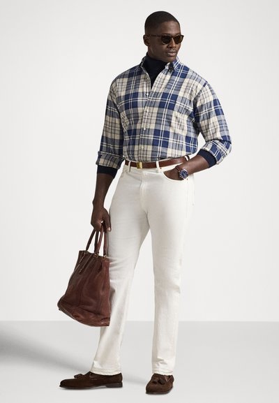 Rutet skjorte i blått og krem, marineblå høyhalset genser, hvite bukser, brun skinnveske, brune loafers med dusker, klokke synlig på håndleddet.