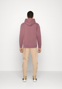 Mężczyzna odwrócony plecami, ubrany w różową bluzę z kapturem, beżowe spodnie cargo i białe sneakersy z zielonymi i purpurowymi akcentami, stojący na tle jednolitej białej ściany.
