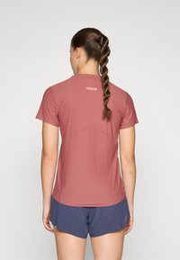 Camisa desportiva de manga curta, rosa, com uma textura suave. Apresenta um pescoço arredondado e o logo HOKA nas costas, combinada com calções azul-marinho.
