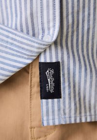 Poignet de chemise rayée bleu et blanc avec une étiquette de marque noire au-dessus de la couture de la poche d'un pantalon beige.