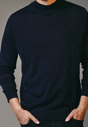 Man met een strakvallende zwarte coltrui, met handen deels in de zakken, tegen een neutrale achtergrond.