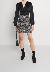 Top negro de manga larga con un escote en V profundo y centro fruncido, combinado con una falda corta de estampado floral con capas. Botines negros y bolso.