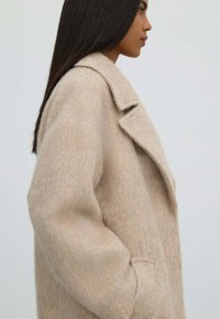 Béžový kabát z vlněné směsi s klopovým límcem, volným střihem a bočními kapsami. Měkká, strukturovaná látka s lehkým vzhledem.