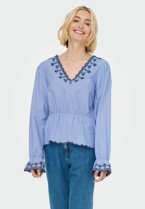 Blouse rayée bleu et blanc avec un col à volants, des détails floraux brodés et des manches évasées, portée avec un jean en denim bleu.