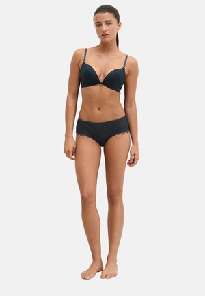 Woman with dark hair tied back wearing black lace bra and matching lace-trimmed boyshorts standing barefoot on a plain light background.