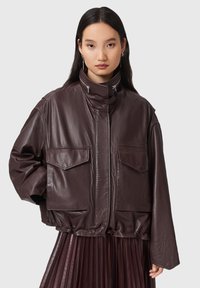 Giovane donna che indossa un giubbotto di pelle marrone scuro oversize con grandi tasche a patta e una gonna plissettata su uno sfondo semplice.