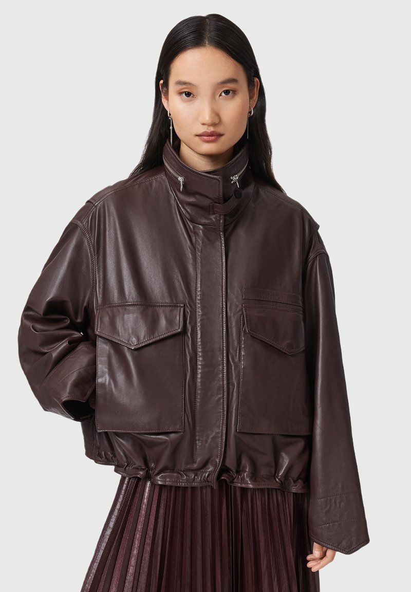 Giovane donna che indossa un giubbotto di pelle marrone scuro oversize con grandi tasche a patta e una gonna plissettata su uno sfondo semplice.