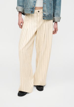 Άτομο που φοράει κρεμ παντελόνι με φαρδιά πόδια και λεπτές μαύρες κάθετες ρίγες, μαύρα παπούτσια, λευκό κοντό τοπ και μπλε τζιν μπουφάν με γυρισμένα μανίκια.