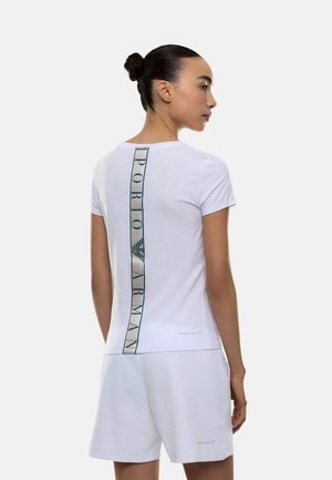 Nainen, jolla on tummat hiukset nutturalla, pukeutuu valkoiseen t-paitaan, jossa on pystysuora "Polo Armani"-teksti ja logo selässä, yhdistettynä valkoisiin shortseihin.