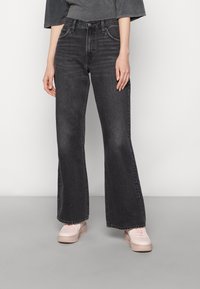 Jean noir taille haute évasés en denim avec une texture délavée. Associés à des baskets rose clair. Vue de face, les mains sur les hanches.