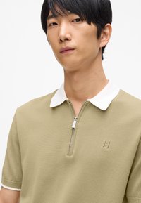 Beige polo-shirt med dragkedja och vit krage, med korta ärmar och en liten broderad logga på bröstet. Slät stickad textur.