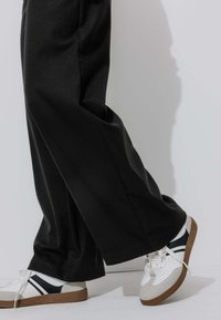 Personne portant un pantalon large noir et des baskets blanches avec des semelles en caoutchouc, marchant contre un fond blanc uni.