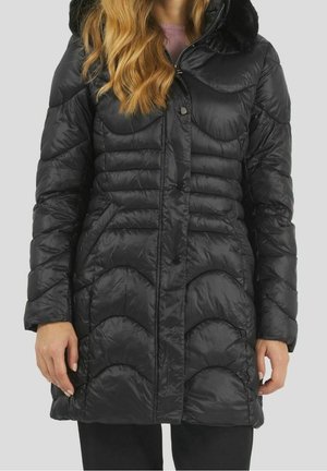 Frau trägt einen knielangen, schwarz gesteppter Steppmantel mit einem pelzbesetzten Kapuzenfutter, steht vor einem schlichten hellen Hintergrund.