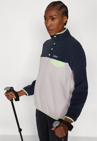 Fleece-pullover med marineblå topp og lys rosa bunn, med en grønn krage og lommeaksenter. Inkluderer knapper og reflekterende detaljer.