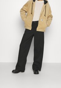 Beige corduroy jas met een zwarte capuchon en ritsen, gecombineerd met zwarte wijde broek en zwarte schoenen. De outfit heeft een ontspannen ontwerp.