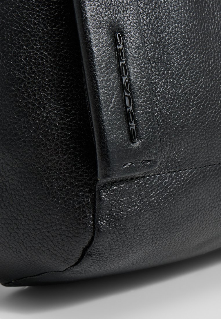 Zwarte leren handtas met een geprofileerde afwerking, een prominent merklogo in zwart en gestikte accenten langs de randen.
